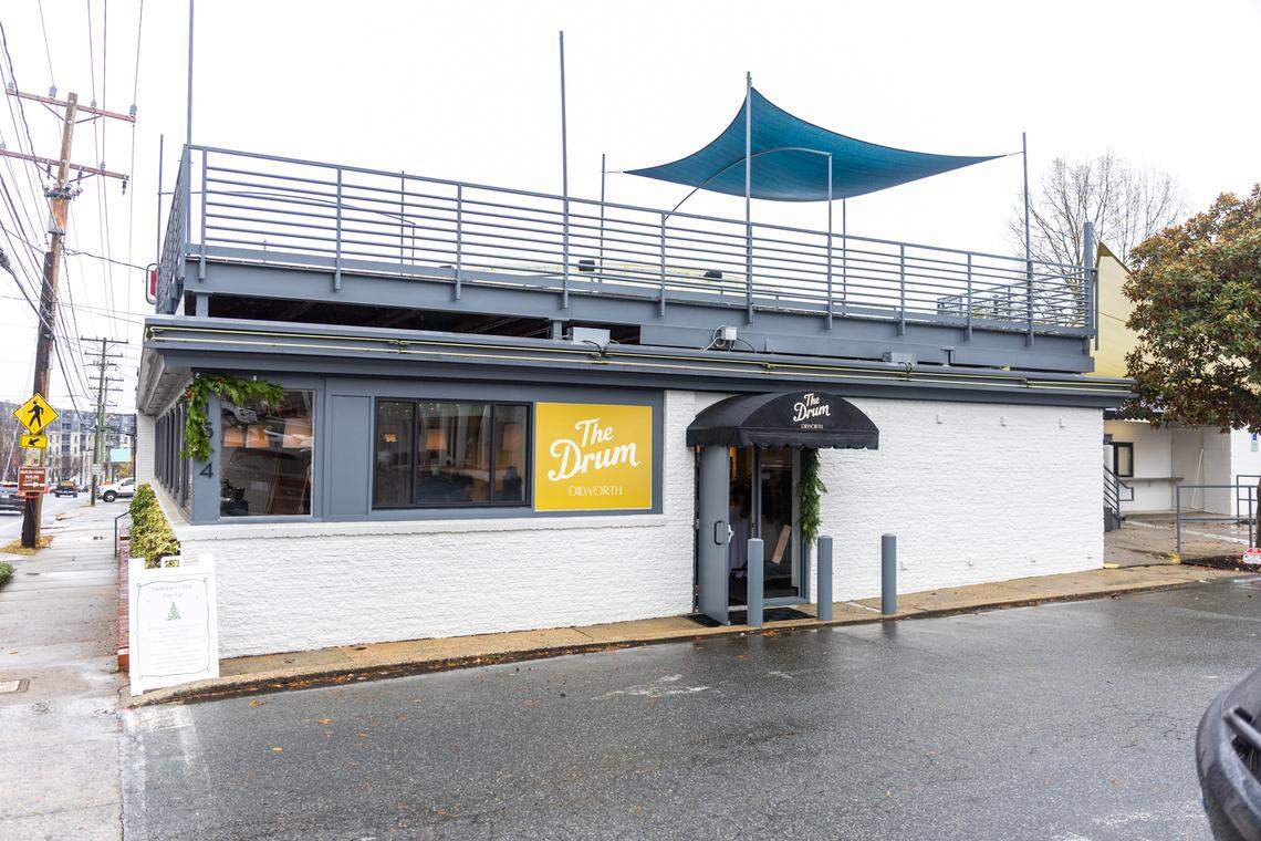 A wide exterior view of “The Drum Dilworth,” a white, textured building with dark gray trim and a rooftop deck with a railing and a dark blue fabric awning. A black, scalloped awning covers the entrance, and an orange square sign reads “The Drum DILWORTH.” An A-frame sign promoting a pop-up market stands near the sidewalk, visible under a cloudy sky.