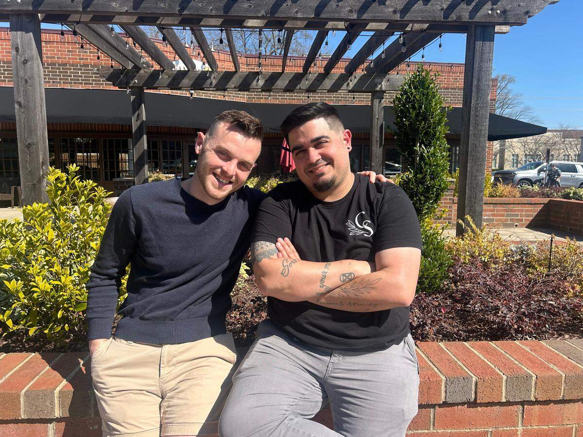 An eye-level, medium shot of two restaurateurs smiling for the camera on a sunny, brick patio. The one on the left, wearing a navy sweater, stands with an arm around the one on the right, who is sitting on a low brick wall wearing a black t-shirt with crossed arms. A wooden pergola, bushes, and a brick building are in the background.