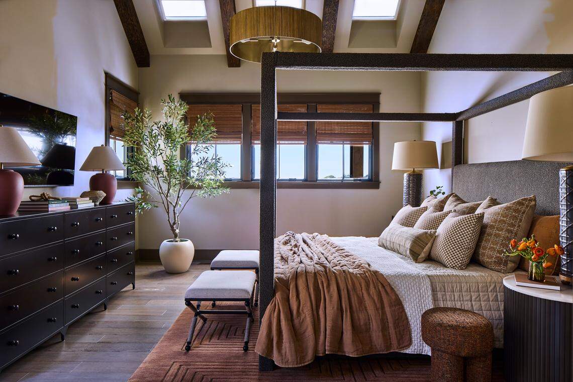 A cozy and luxurious main bedroom with a vaulted ceiling featuring exposed dark wood beams and four small skylights. The centerpiece is a large, dark four-poster bed with textured posts, dressed in white quilted bedding, a russet-colored throw blanket, and numerous textured pillows. A circular, woven gold chandelier hangs over the bed. Two gray upholstered benches sit at the foot of the bed on a large rust-colored rug with a geometric pattern. To the left, a dark brown dresser with silver stud detailing is topped by a television and two reddish lamps. A potted olive tree stands in the corner next to a bank of windows covered with woven bamboo shades.