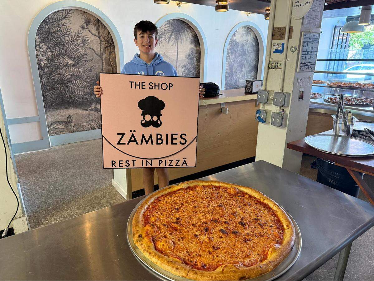An eye-level shot inside a pizzeria featuring a teenager standing behind a stainless steel counter, holding up a massive, square pink pizza box. The large box displays a chef-hatted skull logo along with the words “THE SHOP ZÄMBIES” and “REST IN PIZZÄ.” In the foreground on the counter sits an equally massive, freshly baked New York-style cheese pizza on a round metal tray. The restaurant interior behind him features arched wall niches with tropical, monochrome landscape murals.