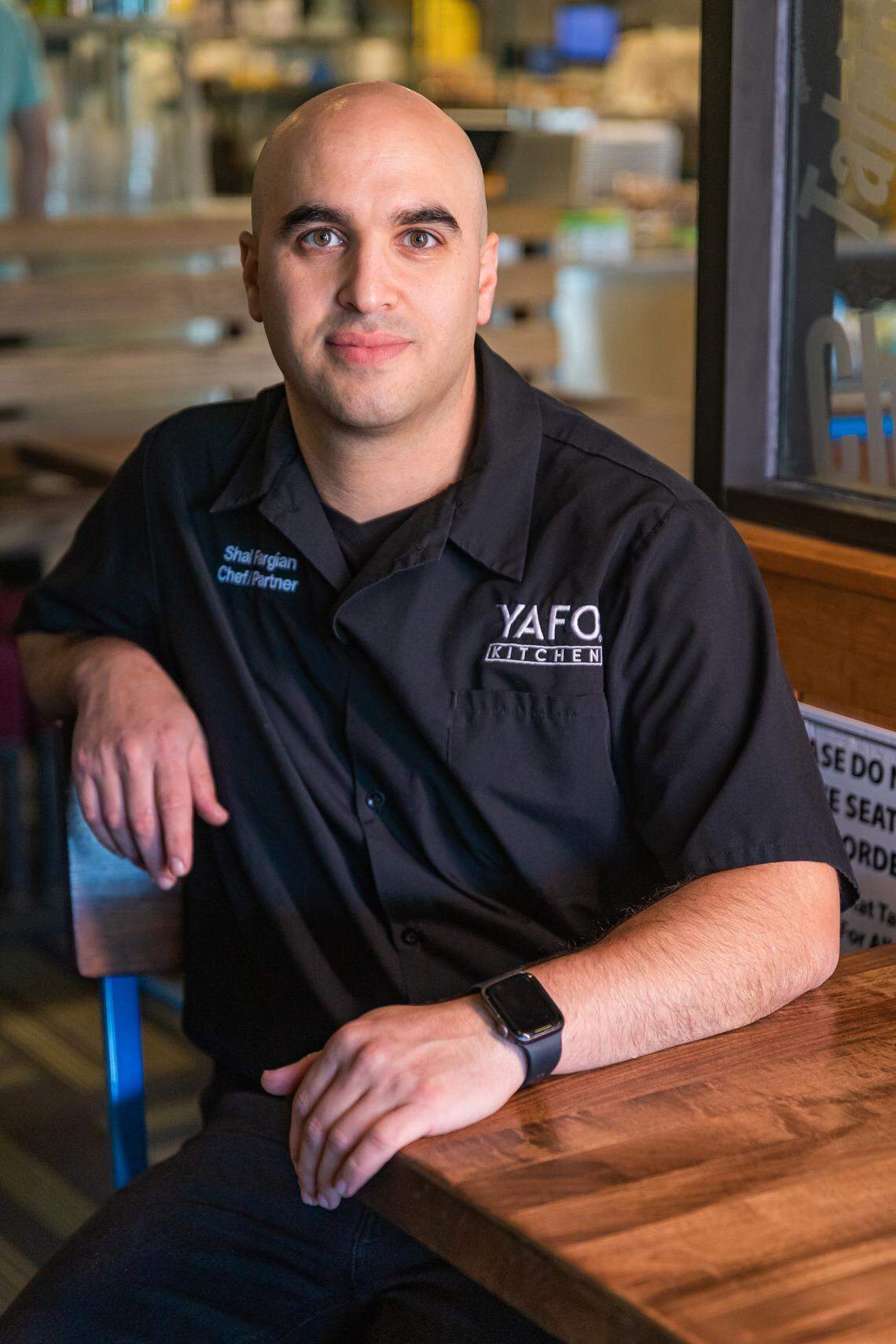 Shai Fargian, Chef/Partner of Yafo Kitchen, poses for a headshot at a wooden table in the restaurant, wearing a black work shirt embroidered with his name and the restaurant’s logo