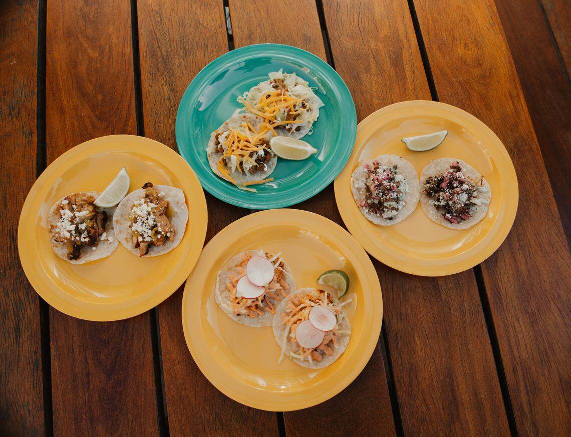 An overhead flat lay shows four plates of tacos arranged on a dark wooden table: three bright yellow plates and one turquoise-green plate. Each plate holds two tacos of varying styles, topped with meat, cheese, radishes, and lettuce, and garnished with a lime wedge.