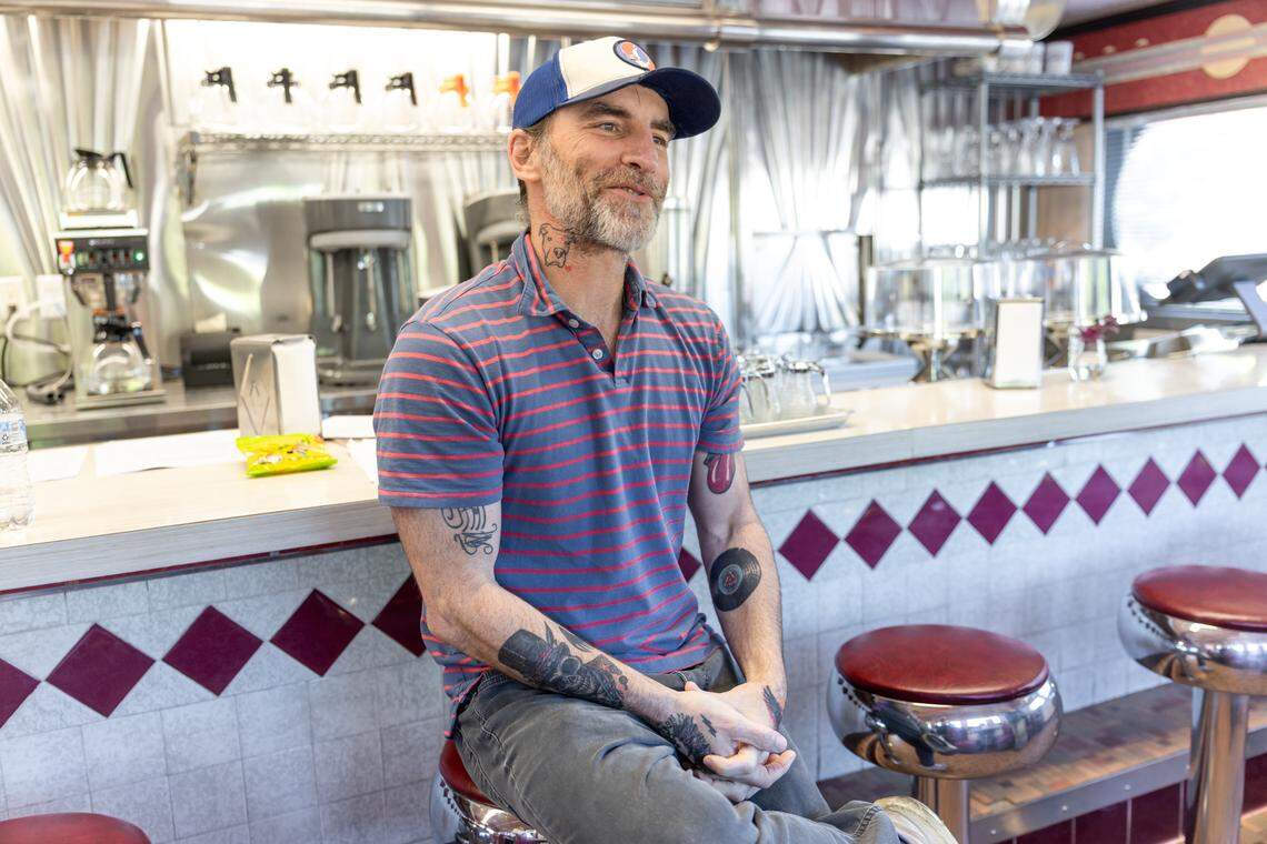 A chef with a grey beard and visible tattoos sits on a red vinyl barstool inside a classic diner, wearing a blue and red striped polo shirt and a blue and white trucker hat, looking off-camera with a slight smile. The background features a vintage-style diner counter with a white tiled front and red diamond accents, along with stainless steel kitchen equipment and coffee makers.