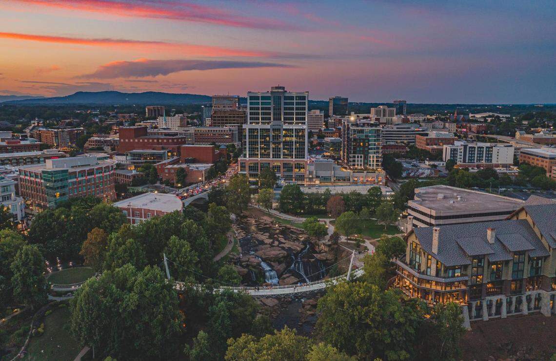 A panoramic aerial photograph of downtown Greenville, South Carolina, during a vibrant sunset. The foreground is dominated by the green space of Falls Park, the rocky cascades of the Reedy River, and the uniquely curved Liberty Bridge. The modern city skyline is lit up in the background, with the Blue Ridge Mountains on the distant horizon.