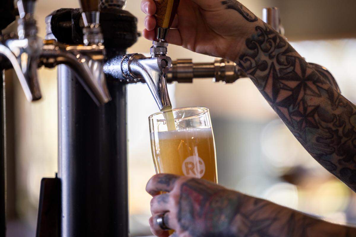 A bartender with tattooed arms fills a clear glass with a light, foamy beer from a silver tap. The glass has a “RC” logo on it. Other taps are visible in the background.