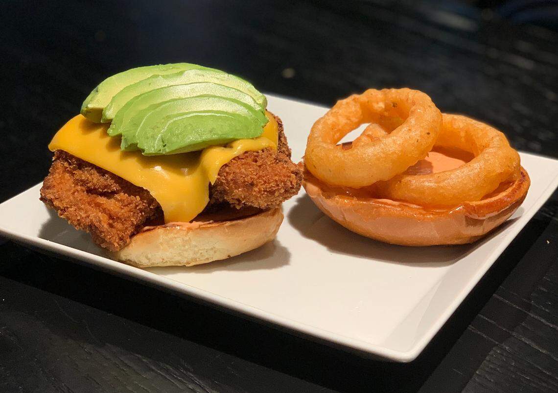 A low-angle close-up shot of a chicken sandwich and onion rings on a white rectangular plate. The chicken sandwich is on a toasted bun and topped with melted cheddar cheese and sliced avocado. The bun for the sandwich is separated, with the top half next to the chicken patty and topped with three golden-brown onion rings. The background is a blurry black table.