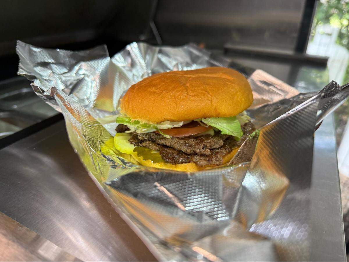 A delicious-looking cheeseburger, wrapped in aluminum foil, is resting on a metallic surface. The burger has a golden-brown bun and is filled with two flat, smashed patties, melted cheese, slices of pickle, lettuce, and a slice of tomato. The foil is partially opened, revealing the burger. The background is slightly blurred.
