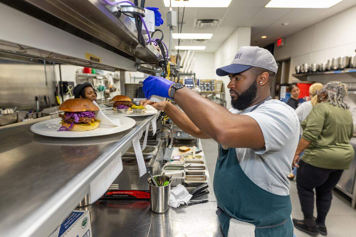 A chef in a light blue shirt, a dark teal apron, and a gray baseball cap is plating a burger in a busy commercial kitchen. The chef, who is wearing blue gloves, is holding a small garnish and placing it on a burger with purple cabbage slaw, which is sitting on a white plate on the metal pass-through shelf. Other kitchen staff and stainless steel equipment are visible in the background.
