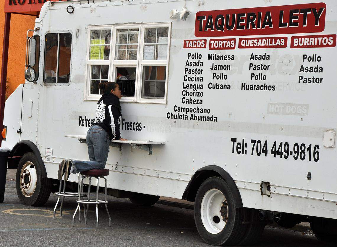 A customer on a stool leans on the counter to order from the Taqueria Lety food truck. The side of the white truck displays a large menu with options for tacos, tortas, and burritos.