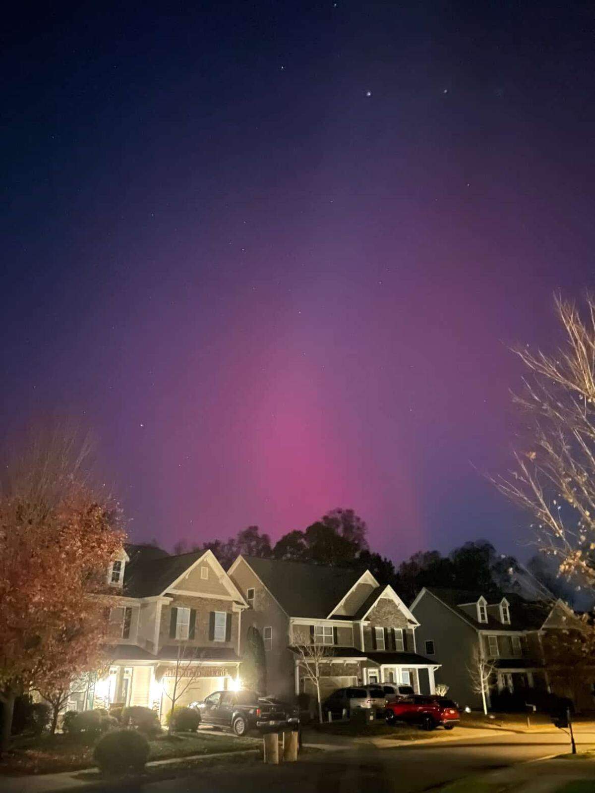 A nighttime photo of the aurora borealis, featuring a wide, deep purple sky with an intense, localized column of bright pink/magenta light rising from the horizon. The foreground is a suburban street with the roofs and facades of several two-story houses visible, some with warm porch lights on. A deciduous tree with autumn leaves is visible on the left.