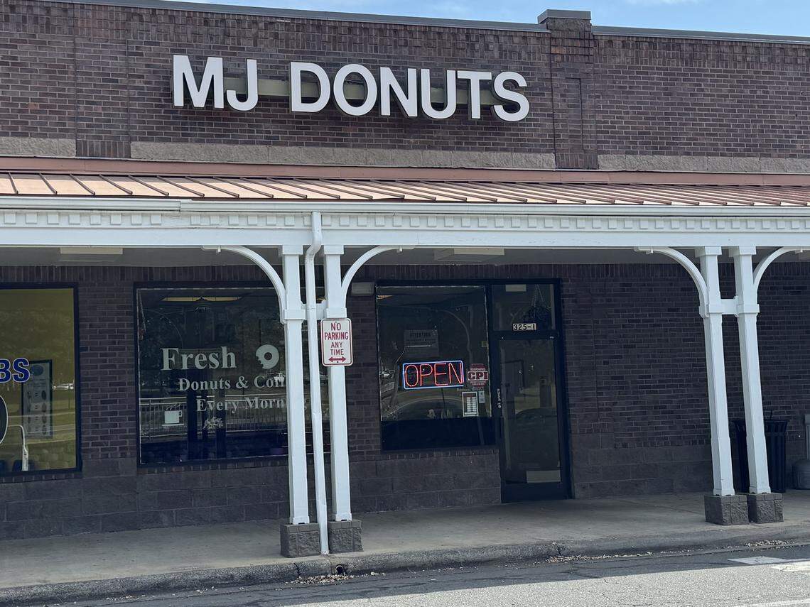 A daytime, street-level view of the entrance to MJ Donuts. The shop is in a brown brick building, with white pillars supporting a covered walkway in front. The name “MJ DONUTS” is in large white letters on the brickwork, and a red neon “OPEN” sign is visible in the door.