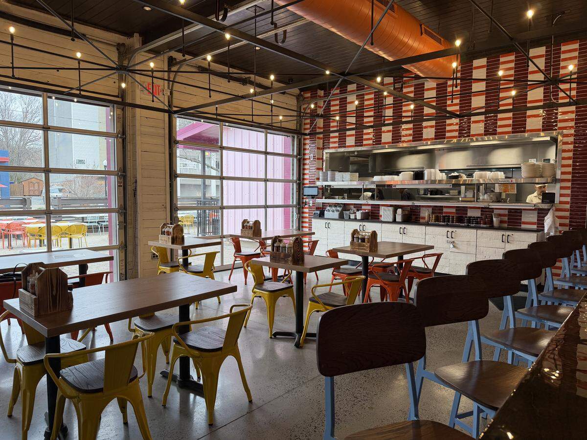 The interior dining room of a restaurant, showing a modern industrial design with high ceilings and exposed orange ventilation pipes. The space features large glass garage-style doors that let in natural light, white brick accents, and a back wall covered in red and white striped tiles behind a stainless steel open kitchen. Square wooden tables are paired with bright yellow and orange metal chairs, while the foreground shows a row of light blue barstools.