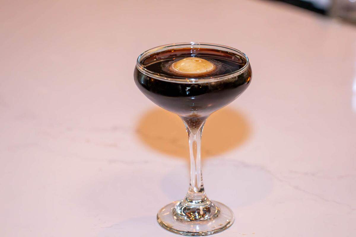 A professional, clean shot of a craft cocktail named “Conspiracy.” The dark, almost black, liquid is served without ice in a stemmed coupe glass. The sole garnish is a small, smooth white ball floating in the middle of the drink. The glass sits on a bright, white marble counter.