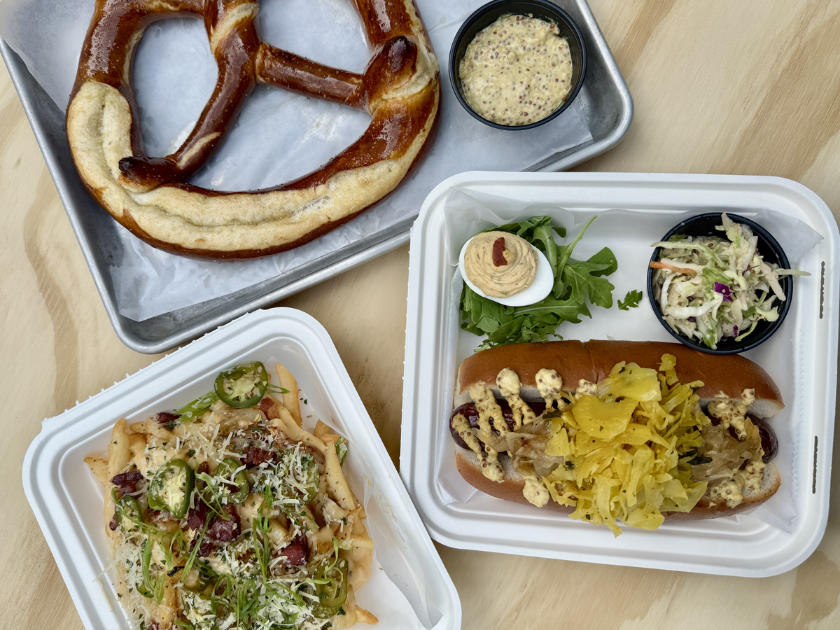 A top-down view of a complete meal spread. In the top left, a large soft pretzel is served with whole-grain mustard. In the bottom left, a container is filled with loaded fries. In the center right, another container holds a bratwurst with sauerkraut, a side of coleslaw, and a deviled egg.