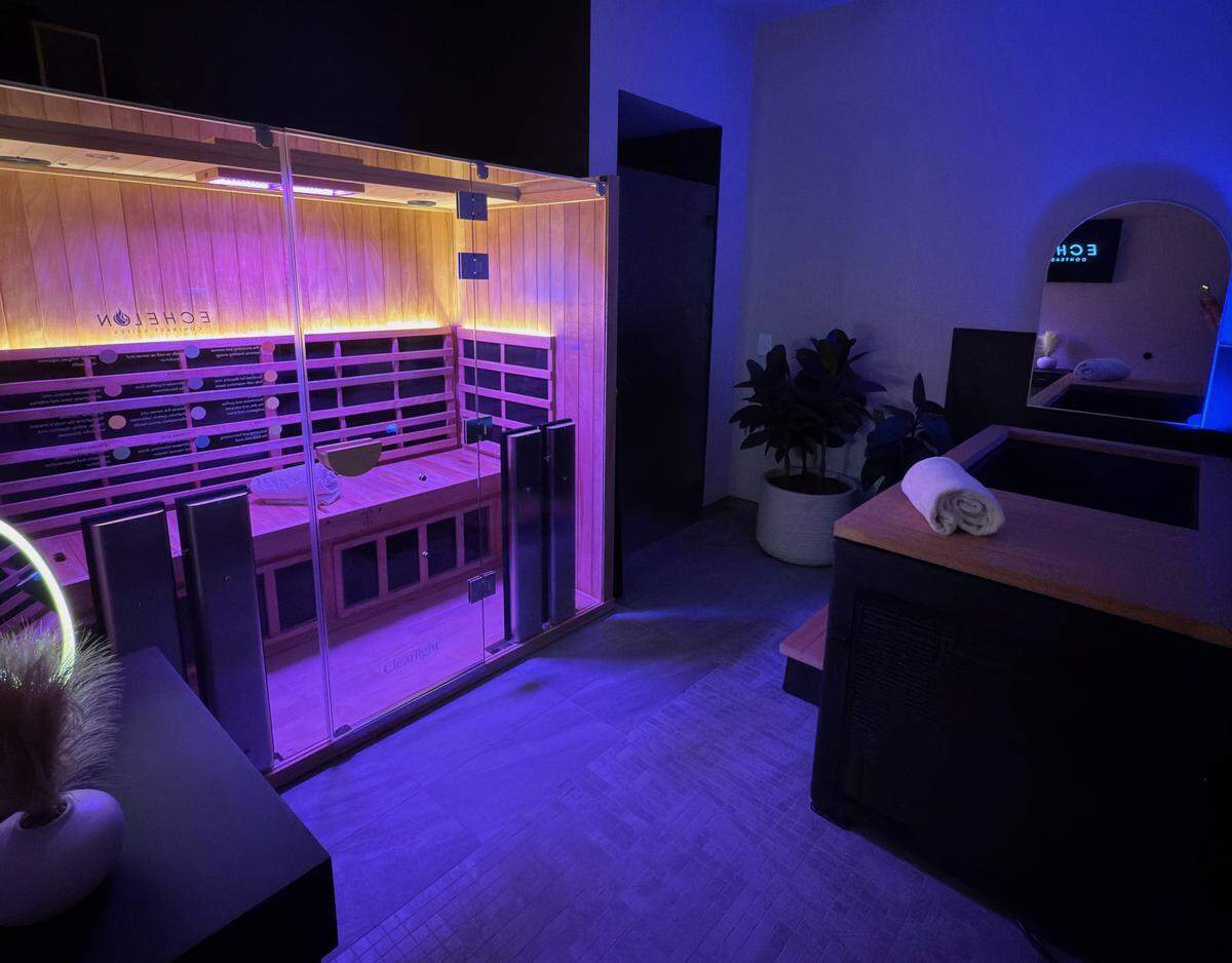 A wide shot of a wellness room bathed in cool blue light. On the left, a large glass-enclosed sauna features light wood paneling, purple-tinted interior lighting, and a logo that reads “ECHELON CONTRAST SUITES.” To the right, a black-paneled cold plunge tub with a wooden top has a white folded towel resting on it. An arched mirror on the wall reflects a digital screen, and a large potted plant sits between the sauna and the tub.