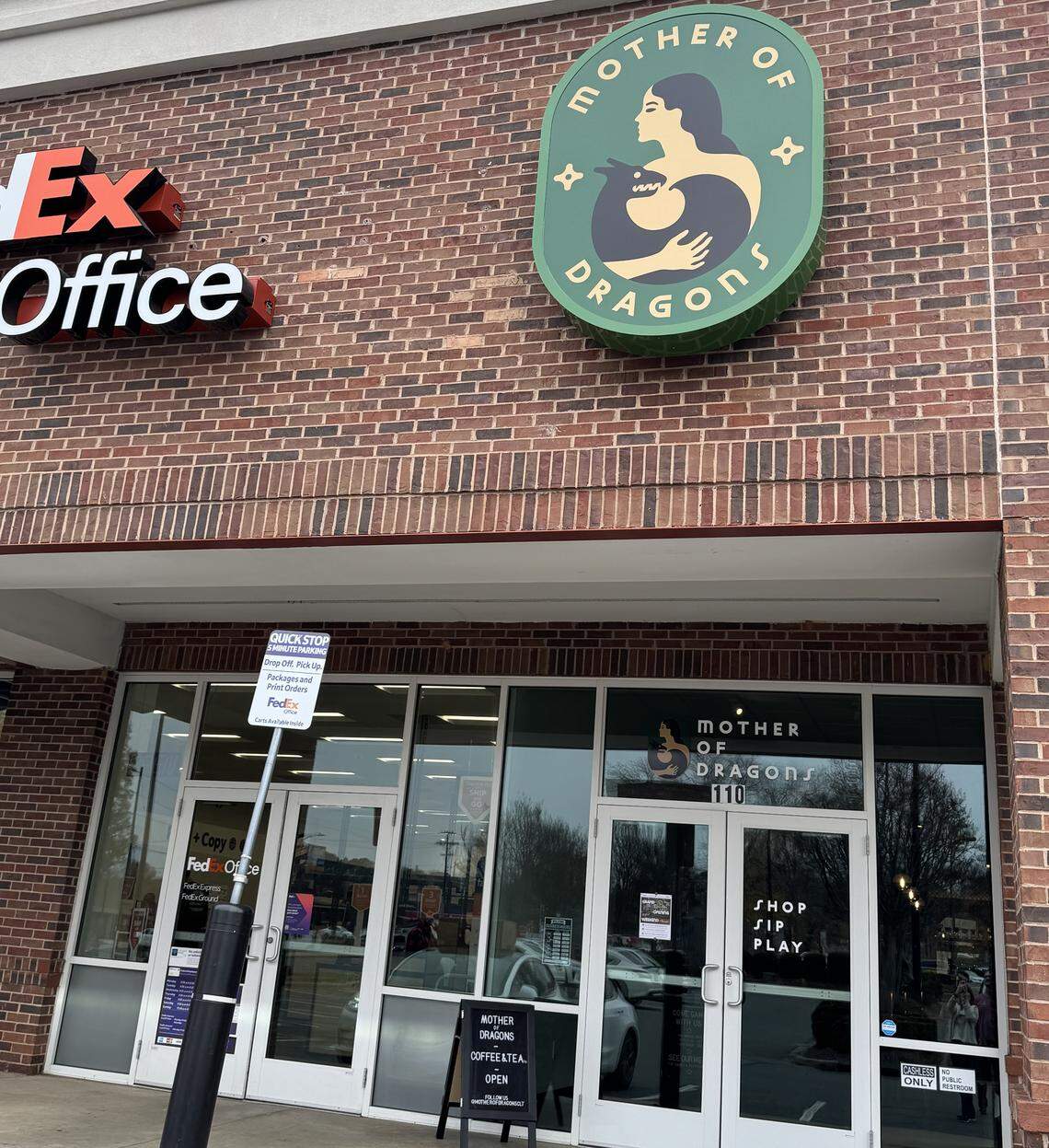 A straight-on, eye-level shot of a red brick commercial building. A large circular green sign with the logo and text “MOTHER OF DRAGONS” is mounted on the brick above the glass storefront. A red, illuminated “FedEx Office” sign is visible to the left.