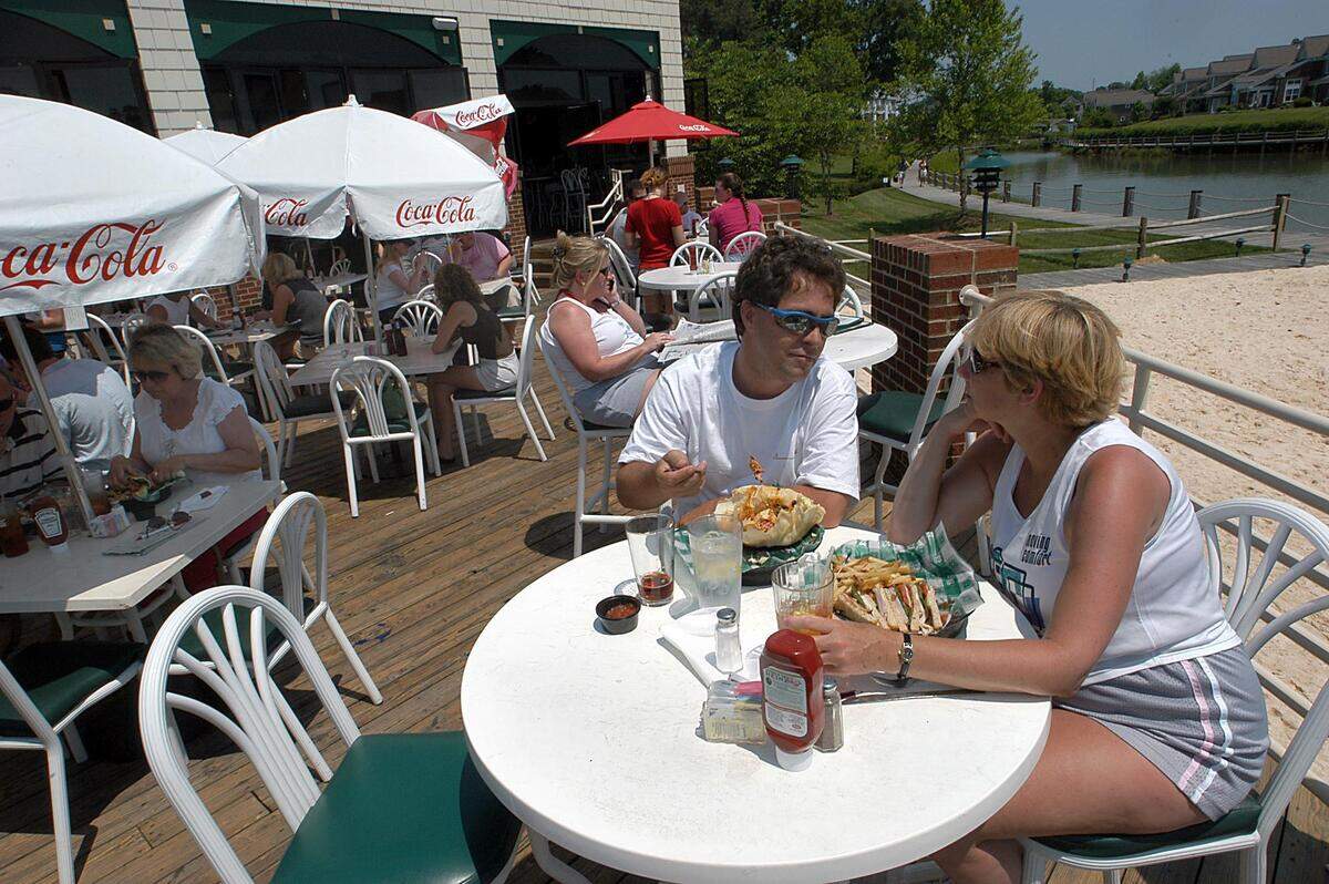 A bustling, sunlit outdoor patio of a restaurant next to a sandy beach and a small lake. Patrons sit at round white tables under white Coca-Cola umbrellas. In the foreground, a man in sunglasses and a white T-shirt and a woman with short blonde hair sit at a table, eating a sandwich with fries and a large taco salad. A wooden walkway and a row of residential townhouses are visible across the water in the background.