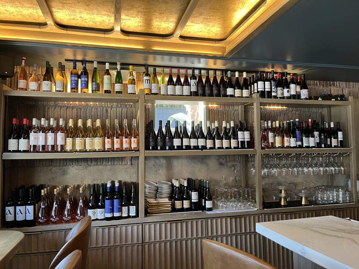 From the perspective of a diner, a large wine wall features multiple tiers of wooden shelves. The shelves are packed with a vast and organized collection of wine bottles. Warm backlighting from the ceiling illuminates the top row. The corner of a white marble table is visible in the foreground.