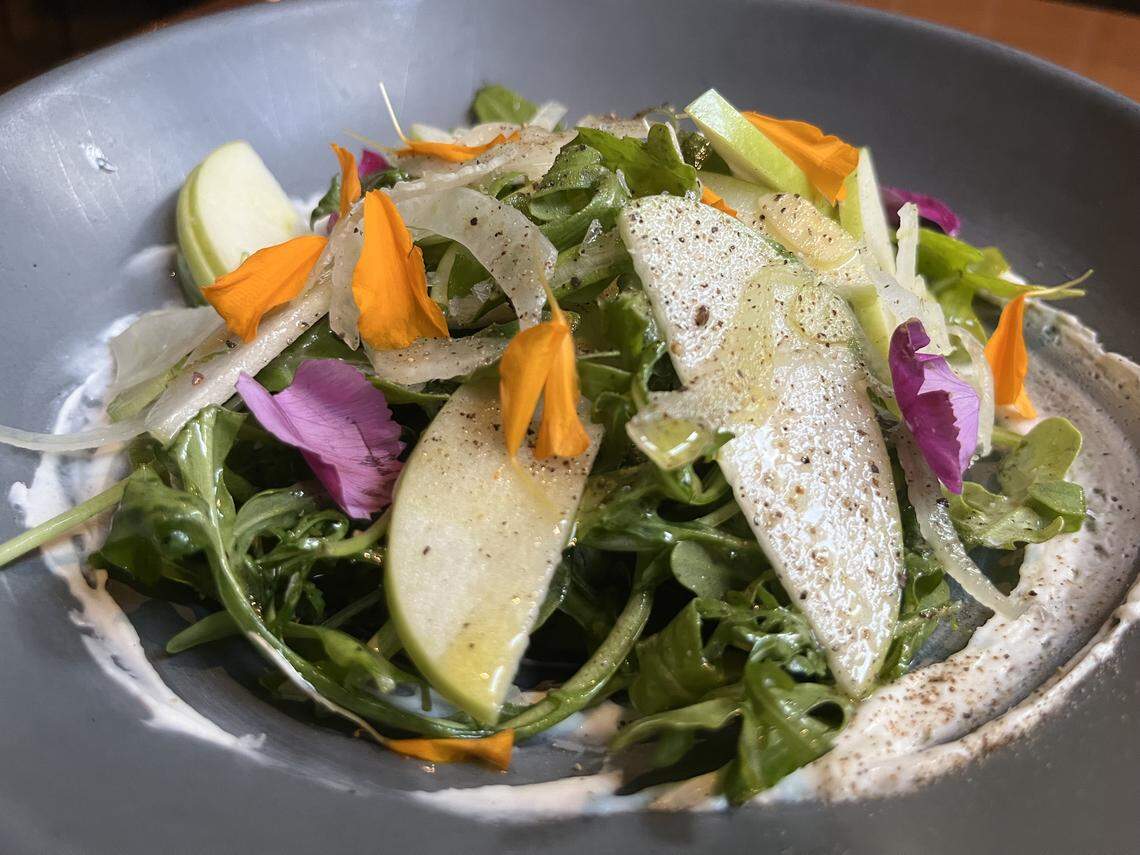 A beautiful, vibrant salad is served in a gray bowl. The salad is a mix of fresh greens, thin slices of green apple, and shaved fennel. It’s dressed in a creamy white sauce that is artfully swirled around the rim of the bowl. The salad is garnished with small orange and purple edible flower petals and a generous dusting of fresh black pepper.