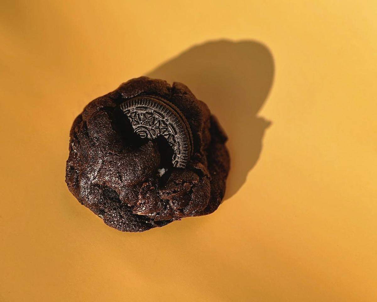 A minimalist studio photograph of a single gourmet cookie. The cookie is a rich, dark chocolate with a fudgy texture, and half of an Oreo is baked into it. It is placed on a flat, mustard-yellow surface under a harsh light that creates a soft, dark shadow.