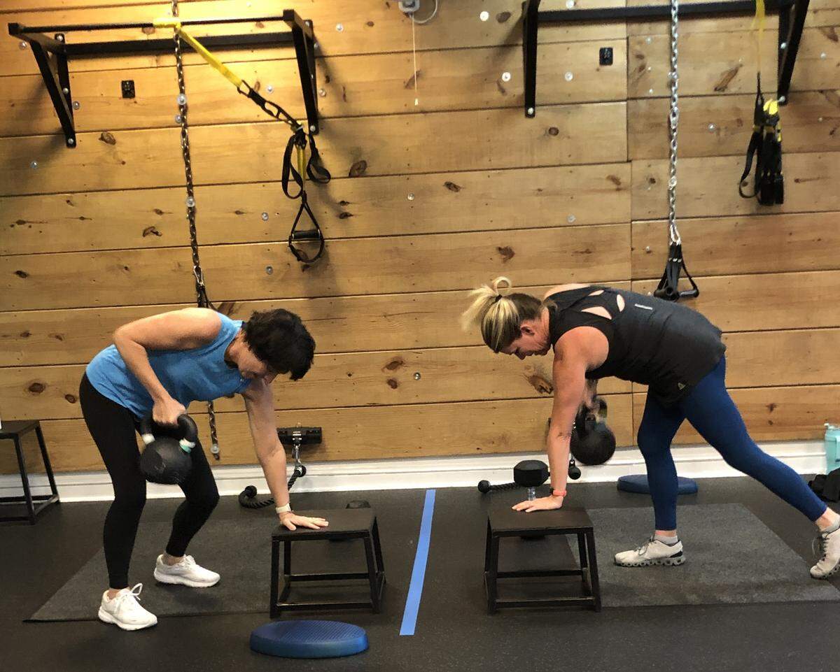 A wide, eye-level shot of two women exercising in a gym with wood-paneled walls. Both women are in a bent-over row position, with one hand on a small black step platform and the other holding a kettlebell. The woman on the left is wearing a blue tank top and black leggings, while the woman on the right wears a black tank top and blue leggings. Suspension trainers hang from the wall in the background.