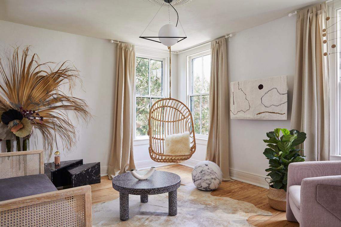 A bright, modern bohemian living room with white walls and wood floors. The center features a wicker hanging chair between two large windows dressed with beige curtains. In the foreground, there is a round black terrazzo coffee table on a light-colored hide rug. The room is decorated with a large dried palm arrangement, abstract wall art, a potted plant, and a light pink upholstered armchair.