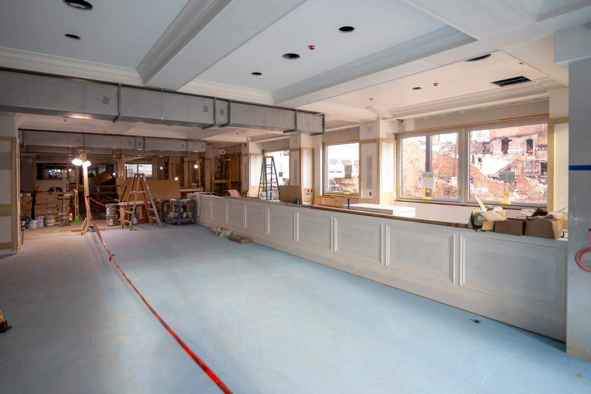 An expansive view of a different section of the interior featuring a long, white-paneled service counter. The floor is covered in blue protective film, and large windows in the background look out toward a brick building ruin.