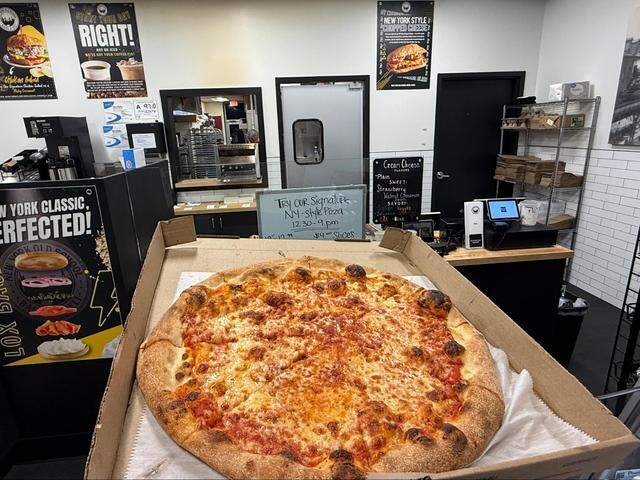 A large, golden-brown cheese pizza with charred crust bubbles sits in an open cardboard box at New York Old School Bagel & Deli. The background shows the shop’s interior, featuring menu signs for New York-style pizza slices and “New York Style Chopped Cheese.”