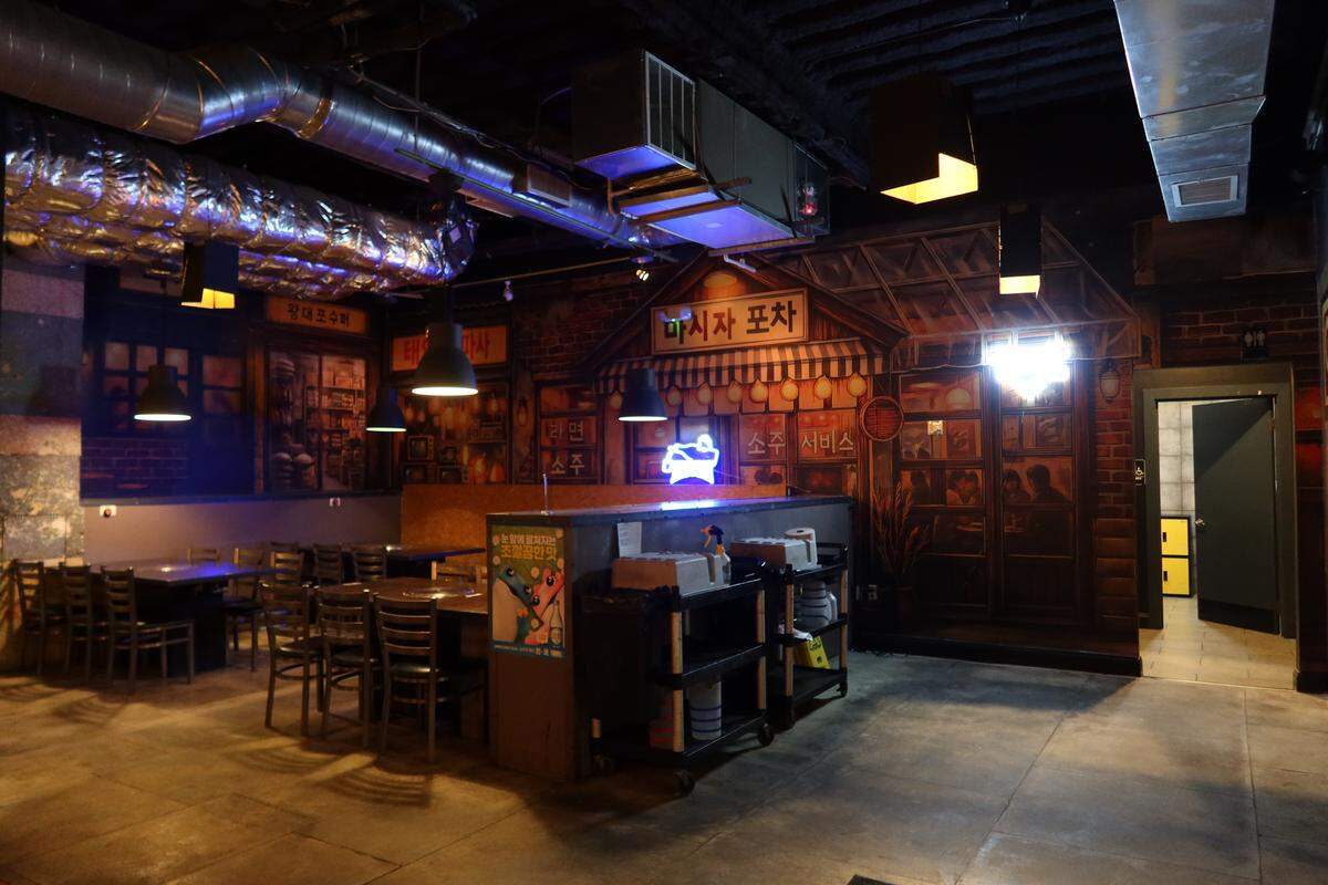 The interior of the restaurant features an industrial design with exposed silver ductwork on the ceiling and a large mural of a traditional Korean street scene. Dark wooden tables and chairs are neatly arranged under dim, warm pendant lighting.