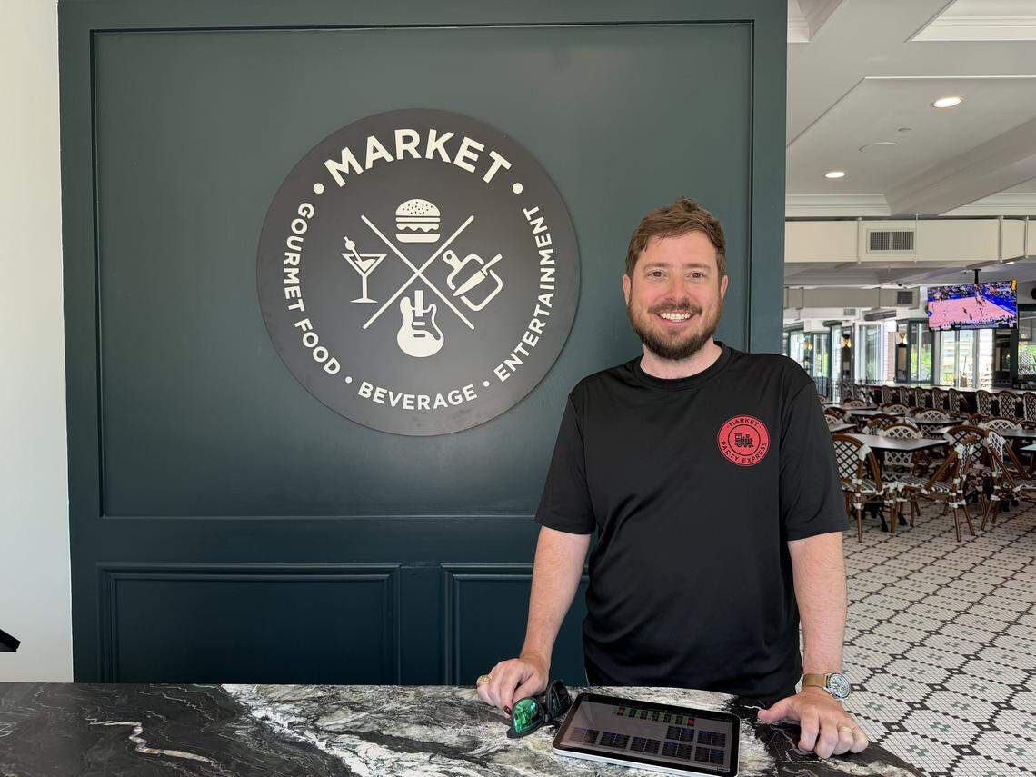 A smiling person in a black t-shirt standing behind a marble counter. Behind him is a dark green wall with a circular “Market” logo featuring icons for gourmet food, beverages, and entertainment.
