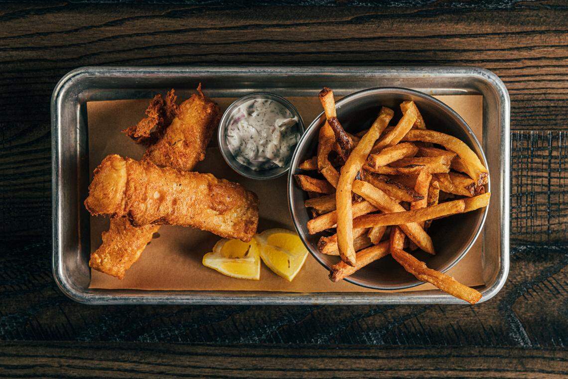 A high-angle shot of a metal tray on a wooden table. On the tray are two large pieces of battered and fried fish next to a bowl of french fries, a small container of tartar sauce, and two lemon wedges.