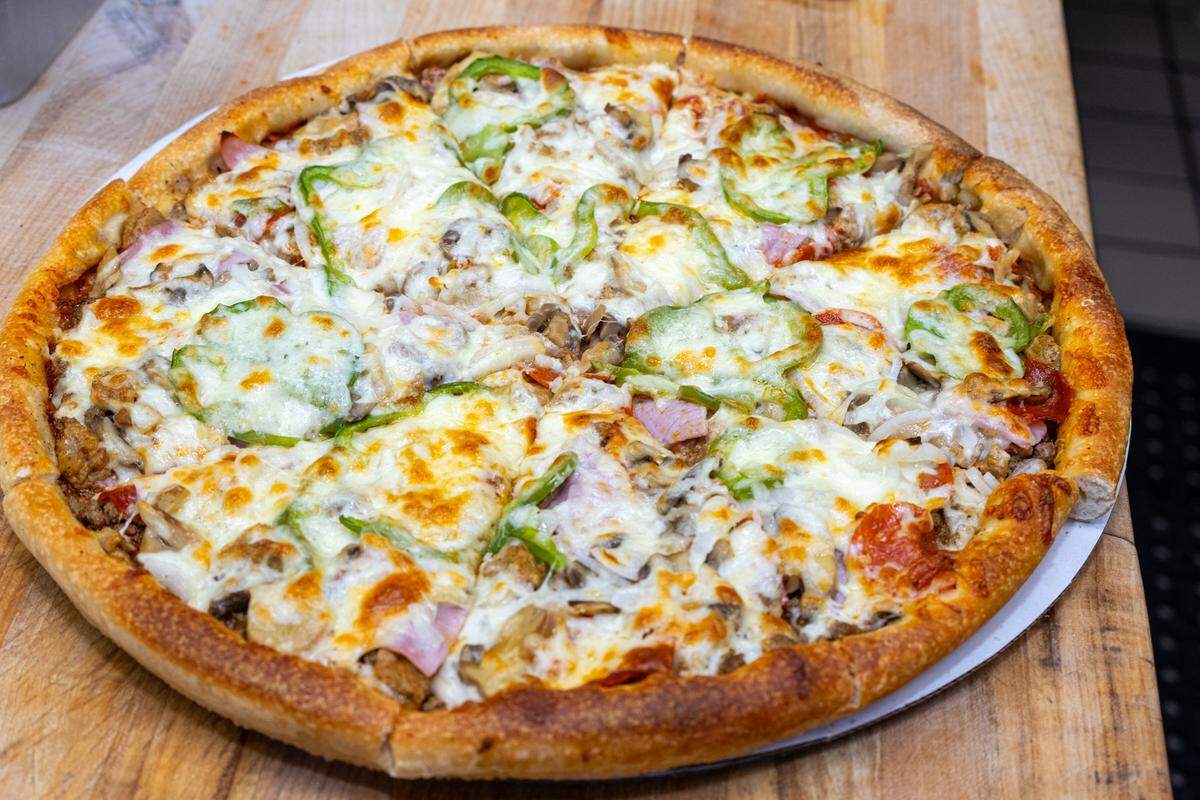 A large, round, freshly baked pizza sits on a light wooden cutting board. The pizza has a golden-brown, thick crust and is generously topped with melted white cheese, slices of green bell peppers, mushrooms, and what appear to be pieces of ham or another meat, along with some red sauce visible beneath the cheese. The cheese has golden-brown bubbly spots, indicating it’s just out of the oven.