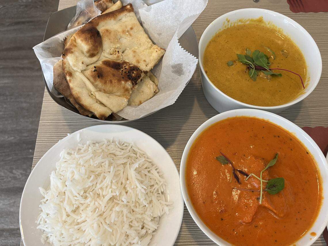 A top-down view of a meal with four components: a metal basket lined with white paper holding pieces of naan bread in the top left, a small plate of white, fluffy Basmati rice in the bottom left, a white bowl of a yellow-orange curry (possibly dal or a vegetable curry) garnished with microgreens in the top right, and a larger white bowl of a bright red-orange curry (likely Butter Chicken or Chicken Tikka Masala) garnished with microgreens in the bottom right. The food is resting on a light-colored wooden table.