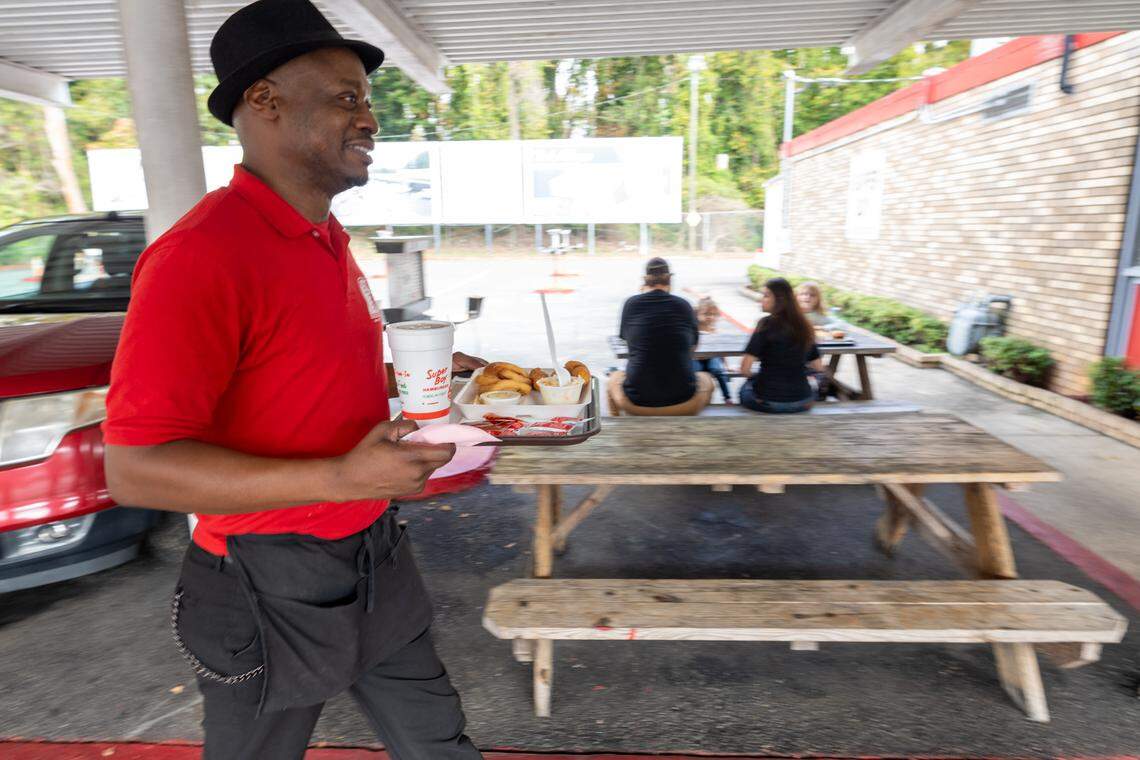A medium, eye-level shot of a smiling restaurant worker, wearing a red polo shirt, black fedora, and a black apron, walking and carrying a food tray. The tray holds a styrofoam cup, onion rings, and a sandwich. The worker is walking through an outdoor seating area with wooden picnic tables. In the background, three patrons are seated at another picnic table, and a large, blurred white billboard is visible beyond a parking lot.