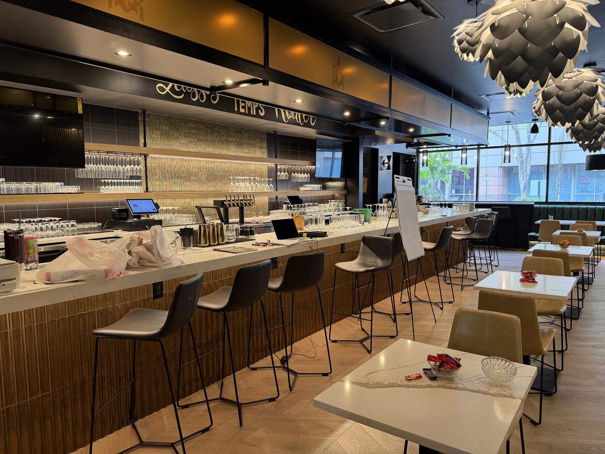 A long, sleek white marble or quartz countertop stretches across the left side of the room. It features a ribbed, golden-wood textured base and is lined with minimalist, high-back grey bar stools with black metal frames. The back wall is covered in shimmering mosaic tiles, lit by an under-shelf warm glow that highlights rows of neatly arranged wine and cocktail glasses. Scripted in gold lettering above the bar is the classic Louisiana phrase, “Laissez Les Bon Temps Rouler” (Let the Good Times Roll), reinforcing the Cajun theme.