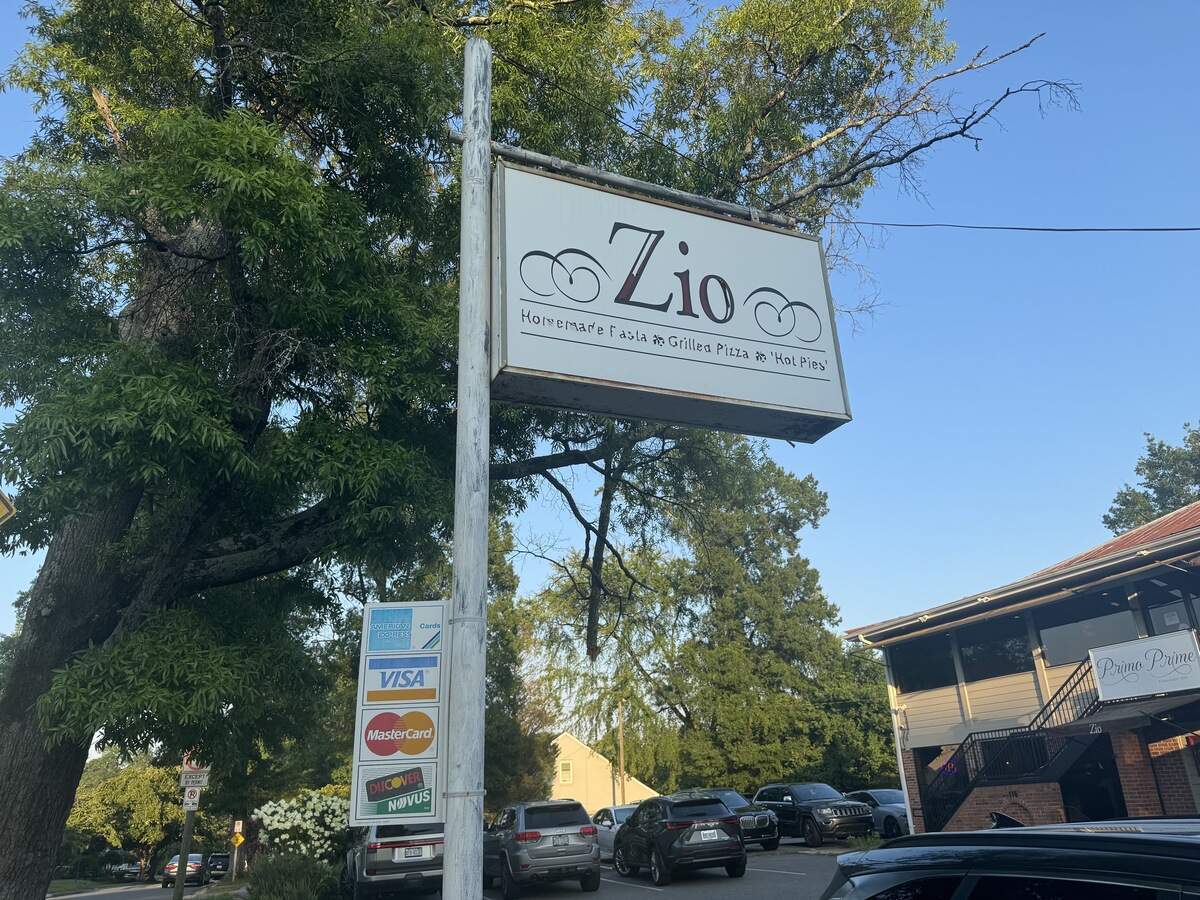 A white, rectangular outdoor sign for a restaurant named “Zio” mounted on a tall metal post next to a mature green tree. Below the large “Zio” logo, the text reads “Homemade Pasta * Grilled Pizza * ‘Hot Pies’”. Lower down on the post, several small credit card logos (American Express, Visa, Mastercard, Discover) are stacked vertically. In the background, cars are parked in a lot in front of a two-story brick and siding building under a clear blue sky.