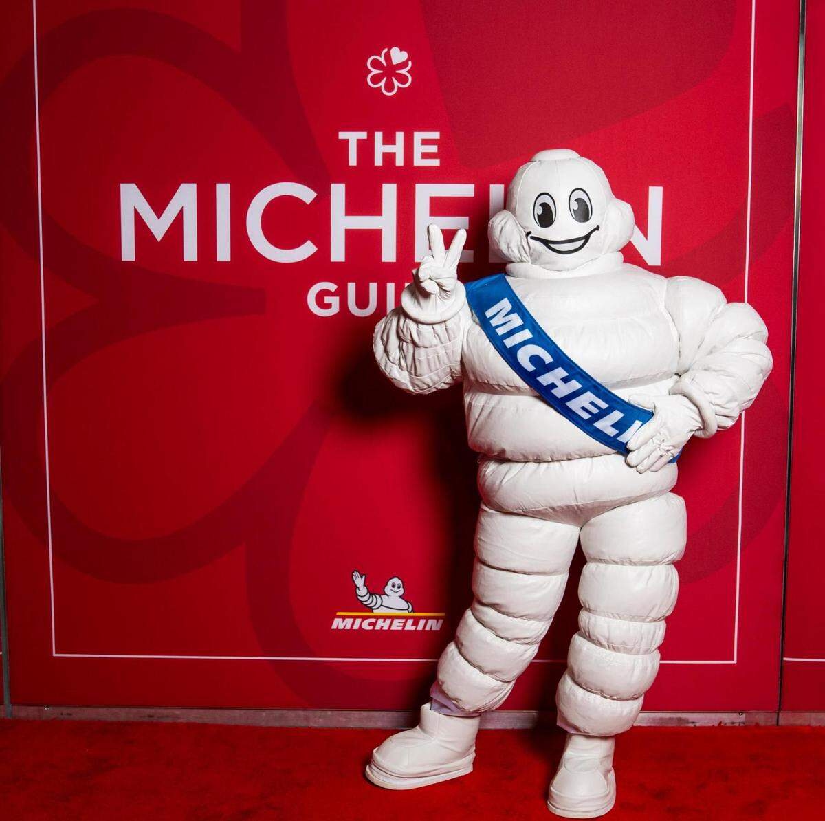 A full-body shot of the Michelin Man character, Bibendum, at a promotional event. He is wearing his signature costume made of white rings and a blue sash. He stands on a red carpet, with one hand on his hip and the other making a peace sign, in front of a red step-and-repeat wall that reads “THE MICHELIN GUIDE.”