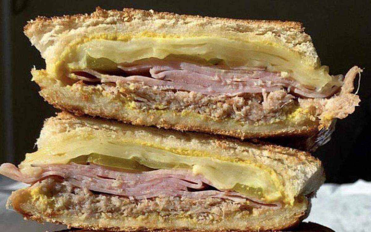 A close-up, cross-section view of a pressed Cuban sandwich cut in half and stacked. The layers include melted cheese, sliced ham, roasted pork, pickles, and yellow mustard on toasted Cuban bread.