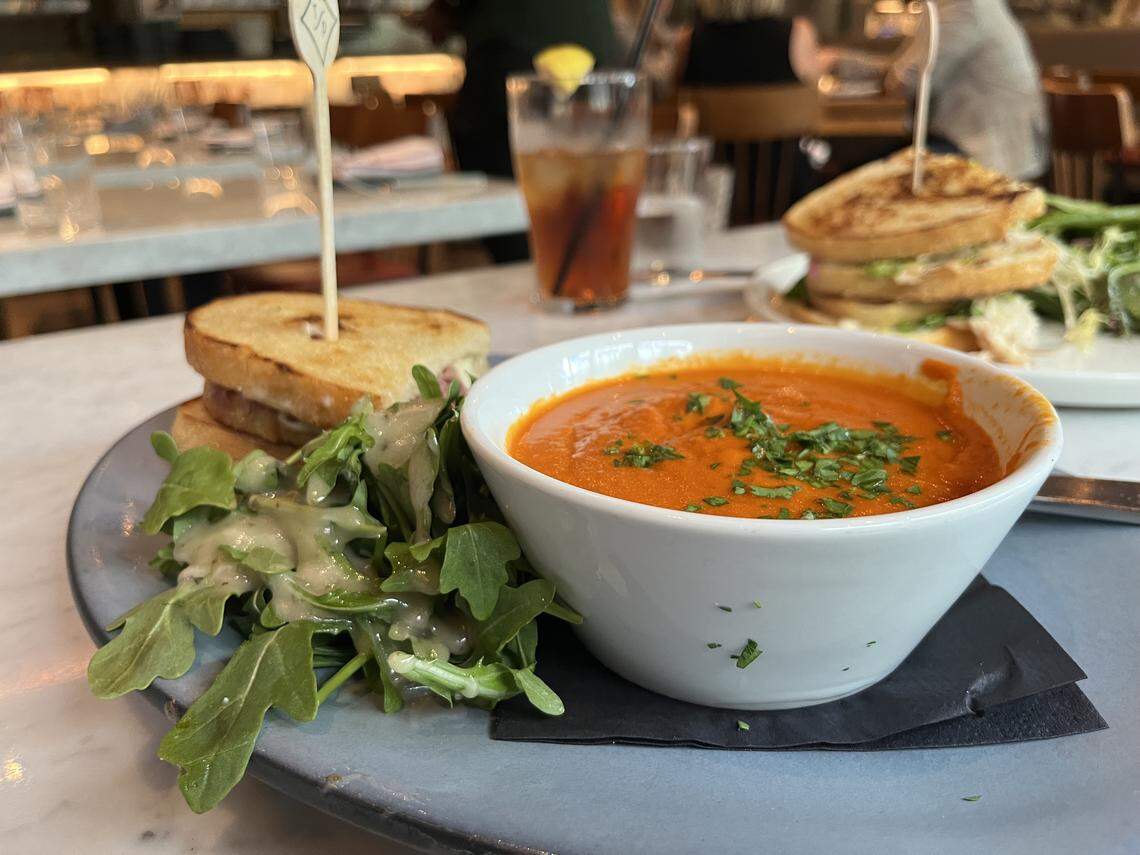 A gray plate holds a bowl of creamy red tomato soup garnished with fresh green herbs. Next to the soup, a half grilled cheese sandwich is served with a small arugula salad drizzled with dressing. Another half of the sandwich is held together by a toothpick. In the blurred background, a tall glass of iced tea with a lemon wedge is visible, along with another sandwich platter and a bustling restaurant setting.