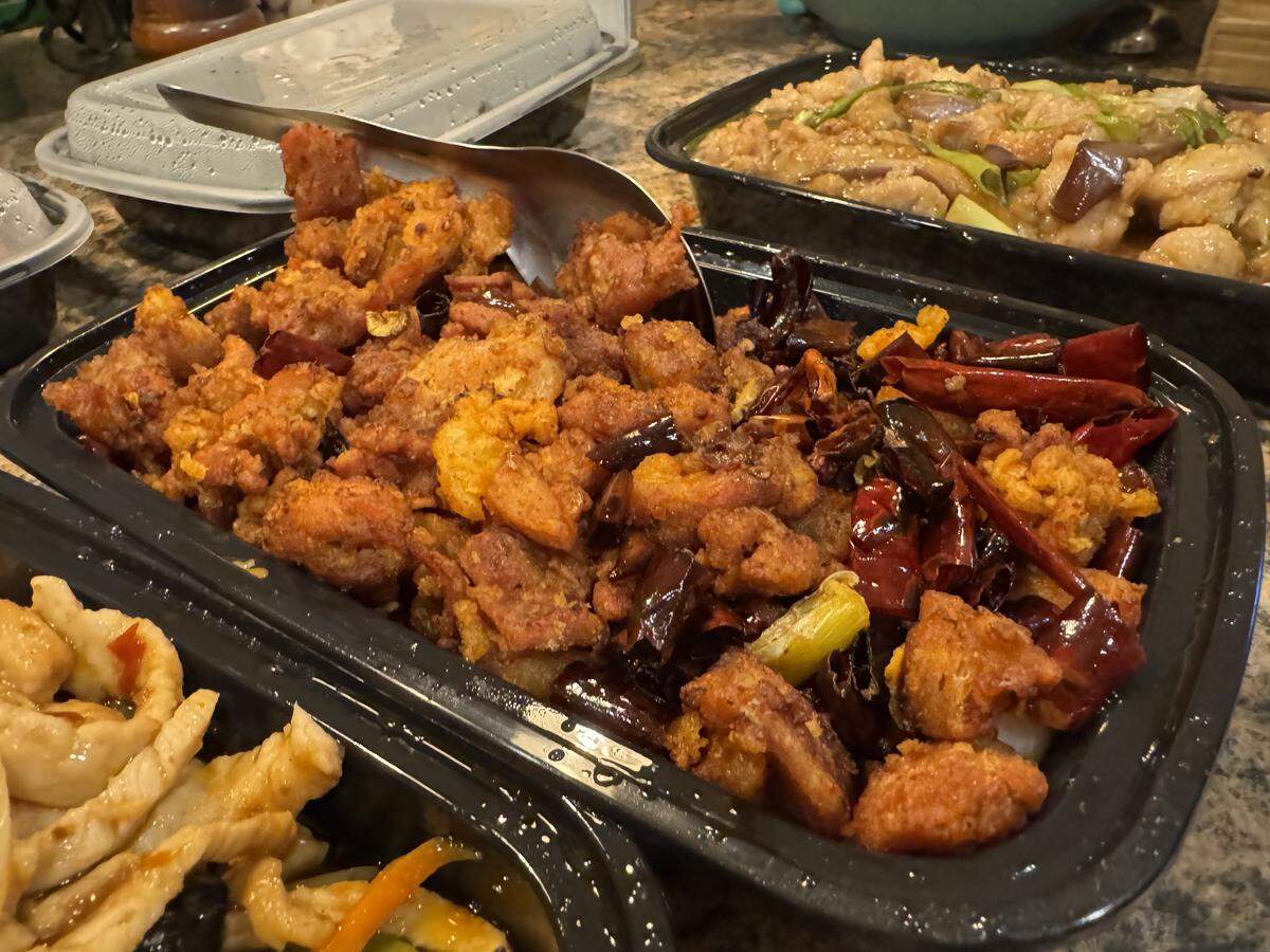 A dish of Dried Chili Chicken from Lee Cafe is presented in a black takeout container as part of a larger meal. The container is filled with golden fried chicken pieces and numerous dried red chilies. A large metal serving spoon rests in the food, and other containers with different stir-fried dishes are arranged on the table.