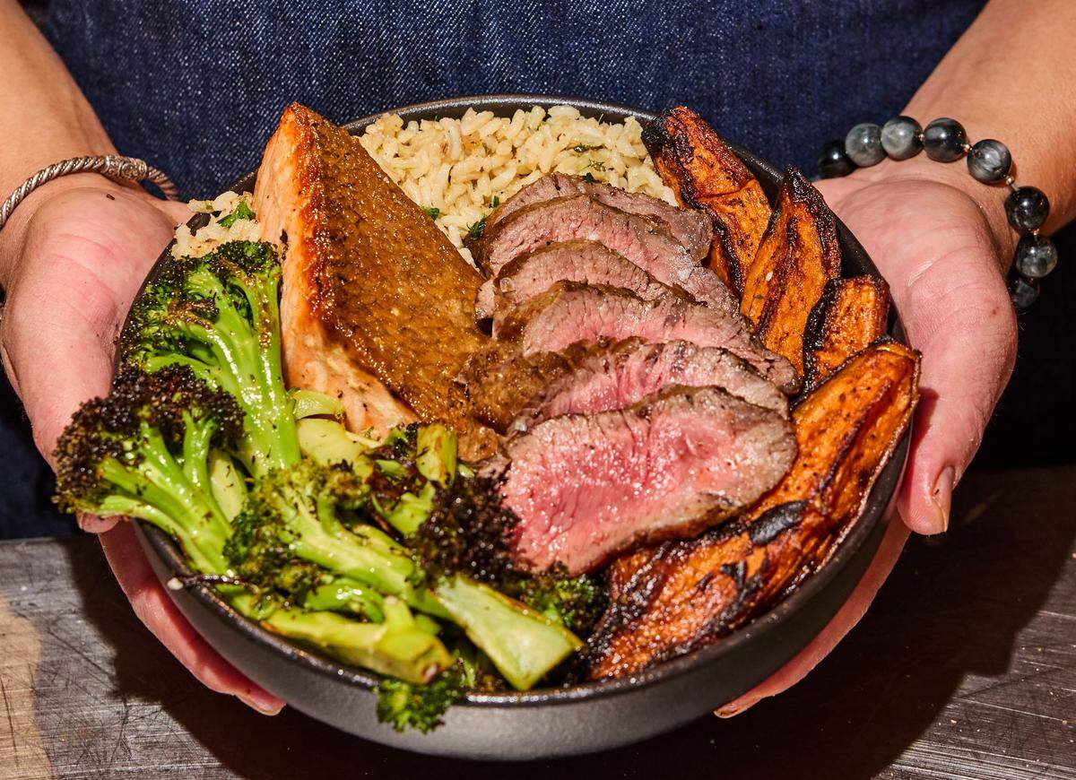 A close-up, eye-level shot of a person’s hands holding a black bowl filled with a hearty, balanced meal. The bowl contains sliced medium-rare steak, a piece of seared salmon, roasted broccoli florets, seasoned rice, and roasted sweet potato wedges. The person holding the bowl is wearing a blue denim garment and jewelry, including a silver twisted bracelet and a beaded bracelet with grey stones.