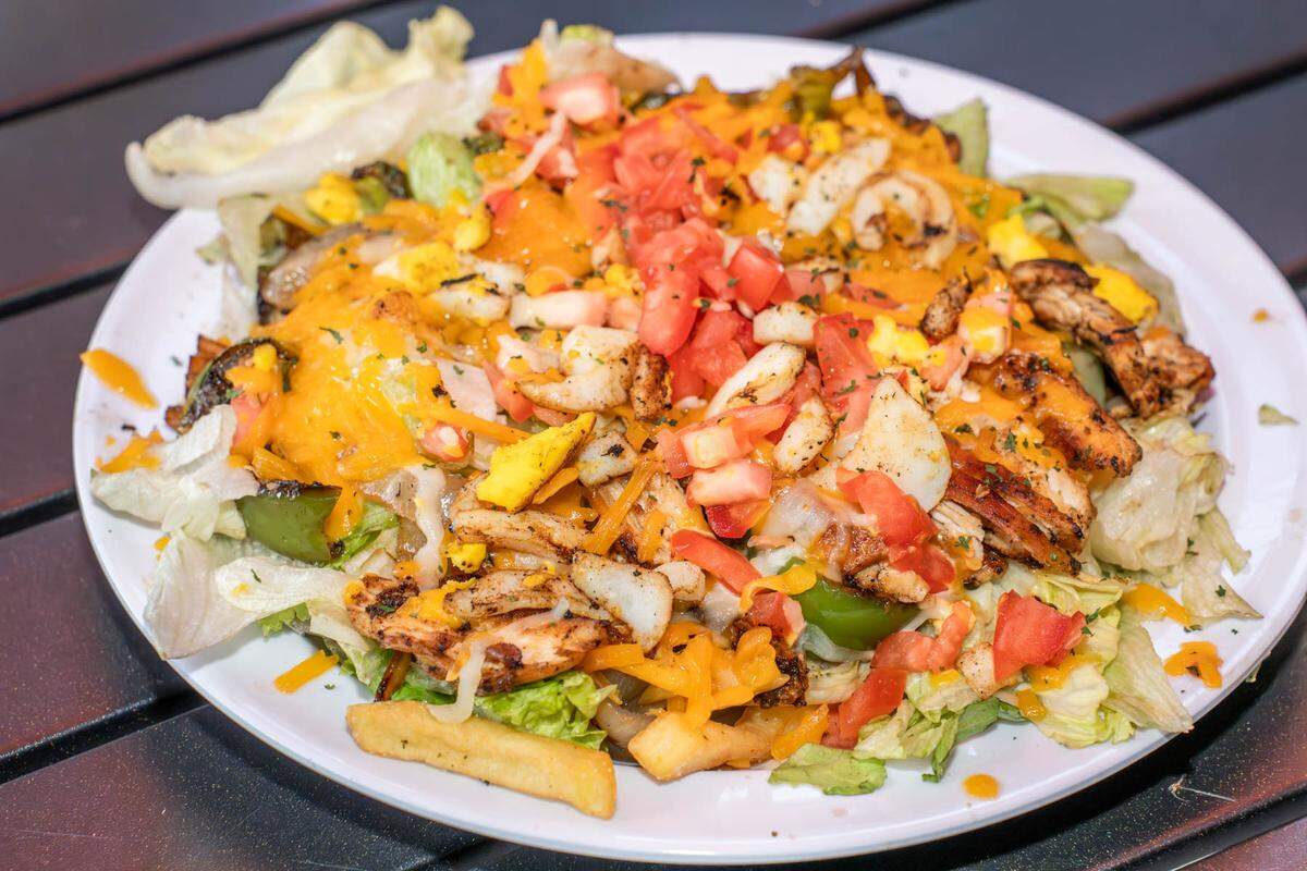 The Sol Salad at Sol’Delish CLT has romaine lettuce on top of french fries with sautéed onions, mushrooms, peppers, tomato, cheese, egg and a choice of protein. This one has chicken with it.