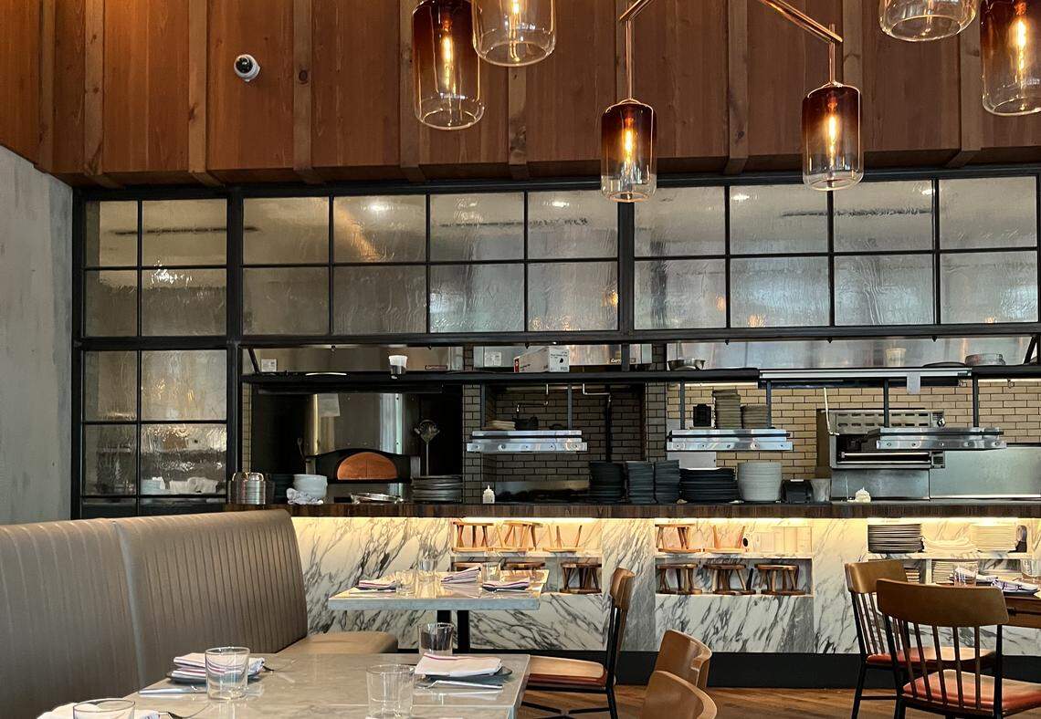 A warm and modern restaurant interior with a view of an open kitchen. The dining area has a tufted banquette seat and tables with white marble-like tops. The open kitchen area features a prominent pizza oven with a tile backsplash, and a counter with stacks of dishes and various cooking equipment. A black-framed window with textured glass separates the dining room from the kitchen. The ceiling is decorated with wood panels and several modern hanging light fixtures with glass shades.