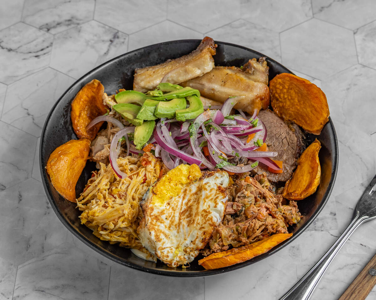 A vibrant and abundant food bowl, photographed from above. The black bowl is artfully packed with sections of juicy shredded chicken, savory shredded beef, crispy-skinned roasted chicken, and a perfectly fried egg. Sliced fresh avocado and tangy pickled red onions top the center, with crunchy sweet potato chips arranged around the edge.