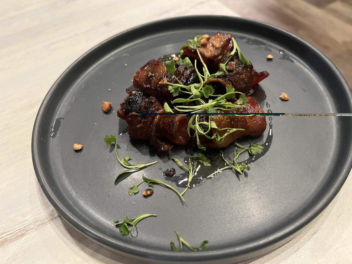 A high-angle, close-up shot of a dark gray, matte-finish plate holding a serving of bacon-wrapped dates. The dates are glazed and caramelized, piled in the center of the plate, and garnished with fresh cilantro microgreens and chopped nuts. The plate rests on a light wood-grain table.