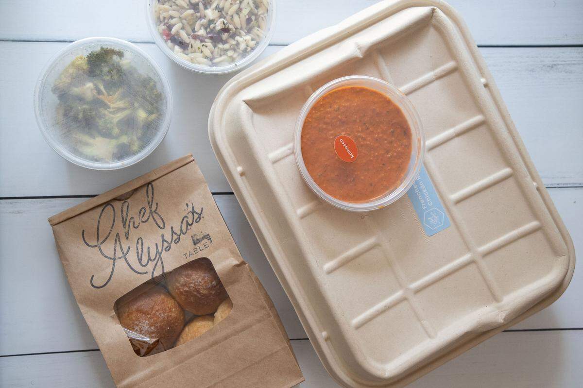 A top-down view of a “Chef Alyssa’s Family Meal” takeout order arranged on a light-colored surface. In the center-right, a large, rectangular, light brown compostable container is topped with a clear plastic container of reddish-orange soup or sauce. To the left, a brown paper bag with a clear window reveals several baked rolls, and “Chef Alyssa’s Table” is printed on the bag. Above the bag, two round clear plastic containers with lids hold side dishes: one with green broccoli and another with what appears to be a grain or pasta salad with red cabbage.