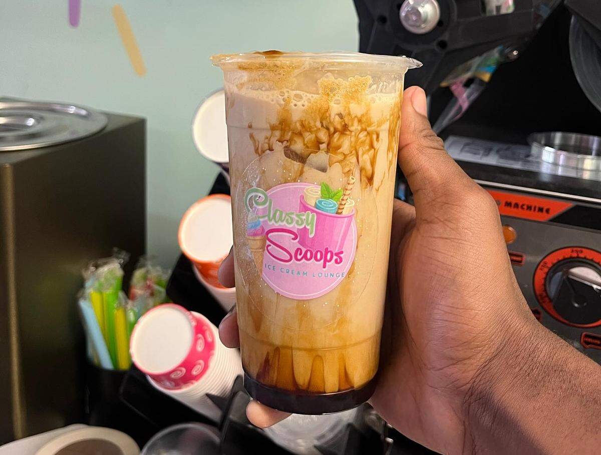 A person holds a tall clear plastic cup of an iced coffee or milk tea that is visibly swirled with dark caramel or brown sugar syrup. The cup displays a “Classy Scoops Ice Cream Lounge” logo and is held in front of cafe equipment.