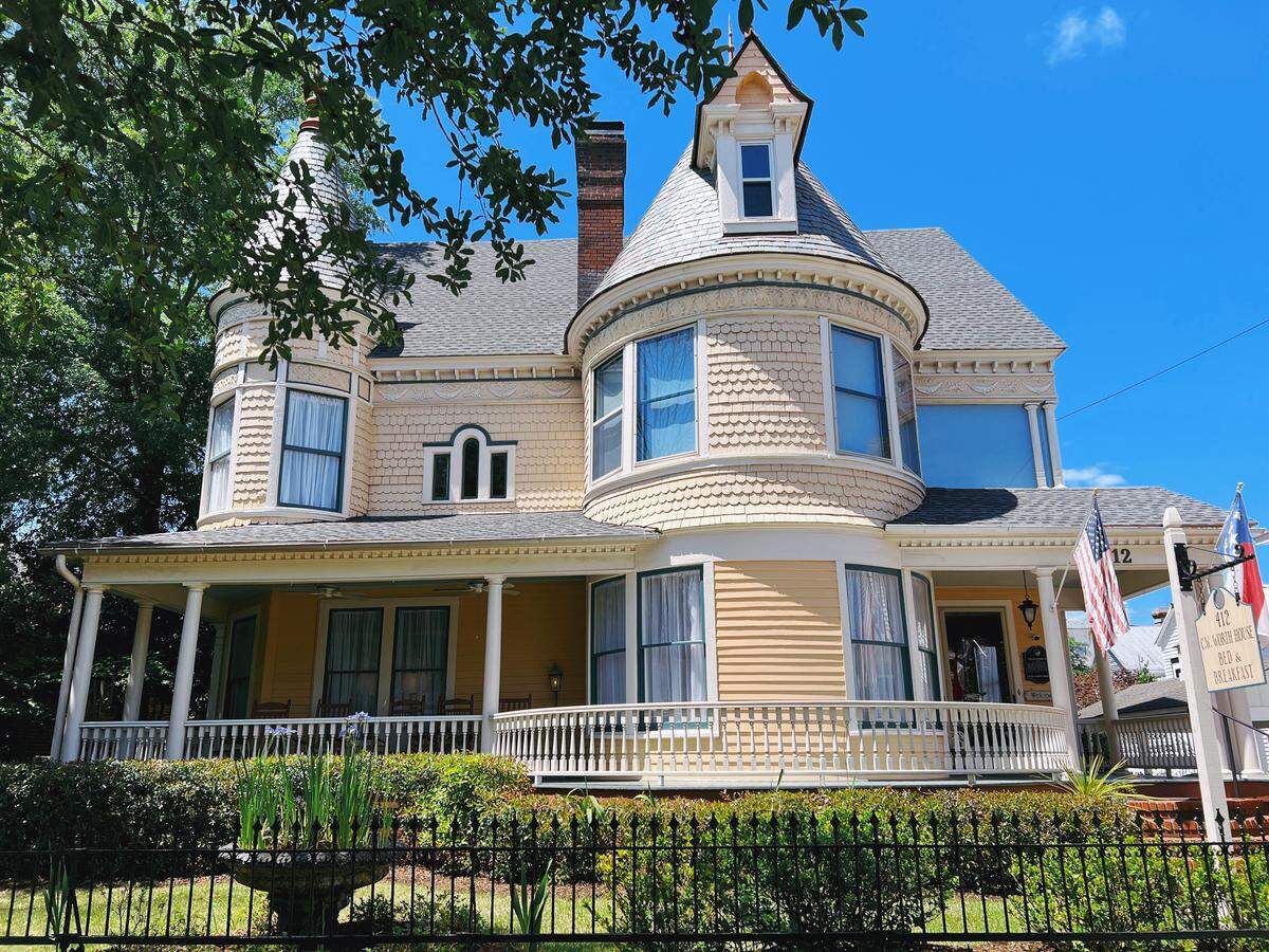 A two-story Victorian house with turrets and a wraparound porch is shown on a sunny day. The house is painted yellow with cream-colored trim and is surrounded by a white fence and lush green landscaping.