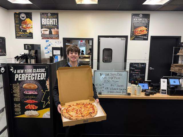 An indoor, eye-level shot shows a person standing behind the wooden service counter, holding open a cardboard box to display a large cheese pizza. A whiteboard advertises “Signature NY-style Pizza” available from 12:30–9 pm, listing prices for whole pies ($12.99–$19.99), $4.00 slices, and a $10 Lunch Special for two slices and a drink. A smaller chalkboard lists Cream Cheese Flavors, categorized into Sweet (Strawberry, Walnut Cinnamon Raisin) and Savory (Scallion, Veggie, Jalapeño, Sun-Dried Tomato, Lox, and Olive).
