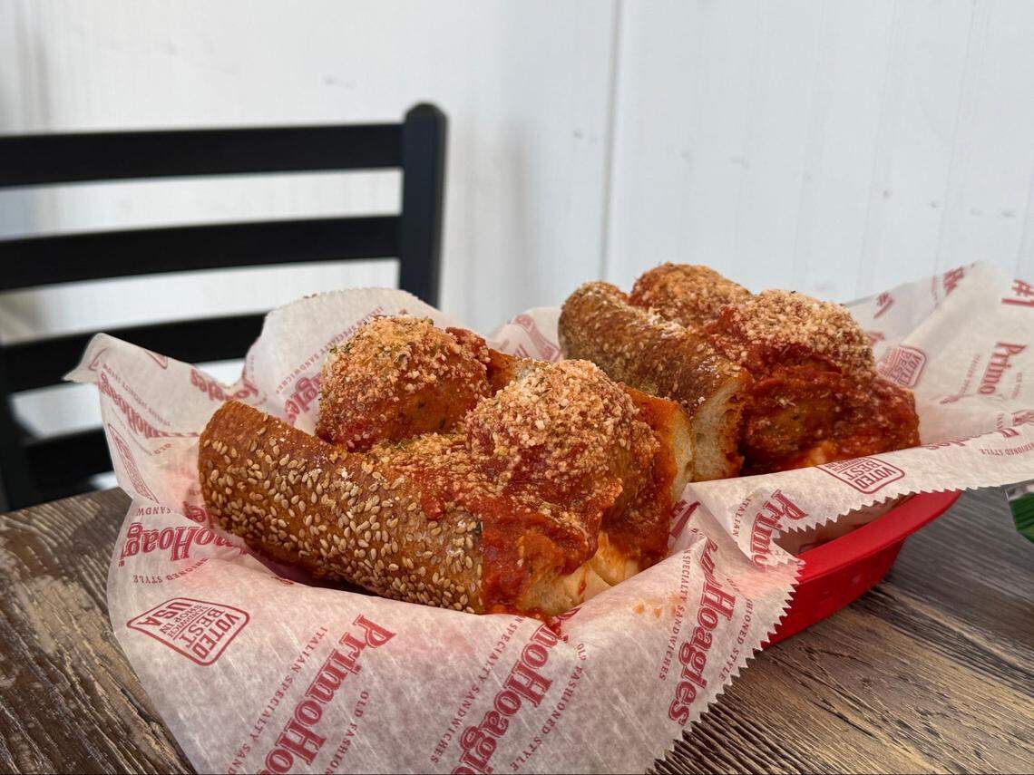 Two large meatball sub sandwiches served in a red plastic basket lined with PrimoHoagies branded wax paper. The meatballs are covered in red marinara sauce and a heavy dusting of parmesan cheese, all held within a crusty sesame seed roll.